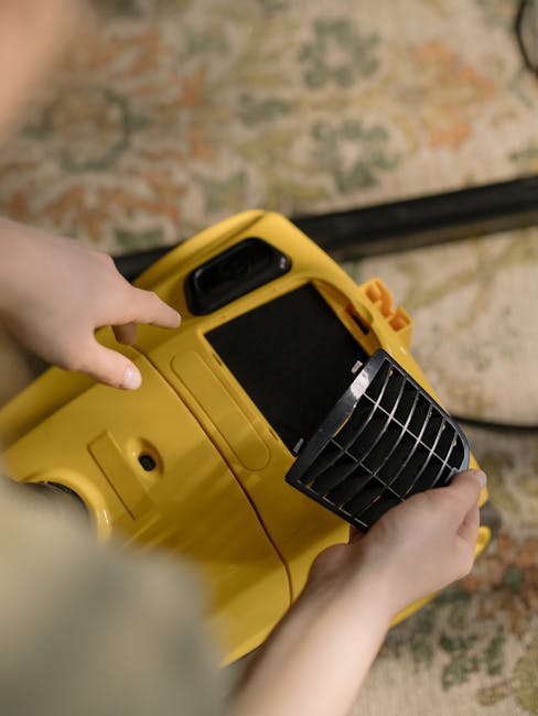 Close-up of a person using a yellow and black vacuum cleaner on a patterned carpet inside a residential room. The individual, wearing blue jeans and a neutral-colored top, is holding a cloth within th