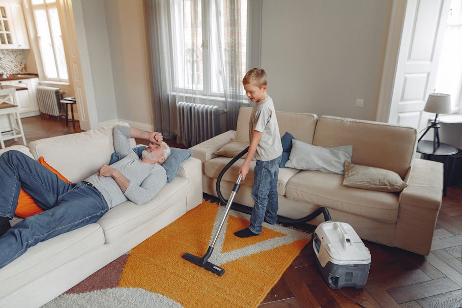 A young boy using a vacuum cleaner to perform surface cleaning on a beige sofa in a living room, while an elderly man lies relaxed on the same sofa. The room features wooden flooring partially covered by a yellow and gray rug, light gray walls, and large windows with sheer curtains allowing natural light. A side table with a lamp is visible next to the sofa. The vacuum cleaner, with a hose attachment, is positioned on the floor nearby, indicating ongoing deep cleaning and hygiene maintenance as part of a domestic cleaning process, with a focus on keeping furniture clean and fresh. The scene illustrates routine household cleaning in a well-lit, tidy living space.