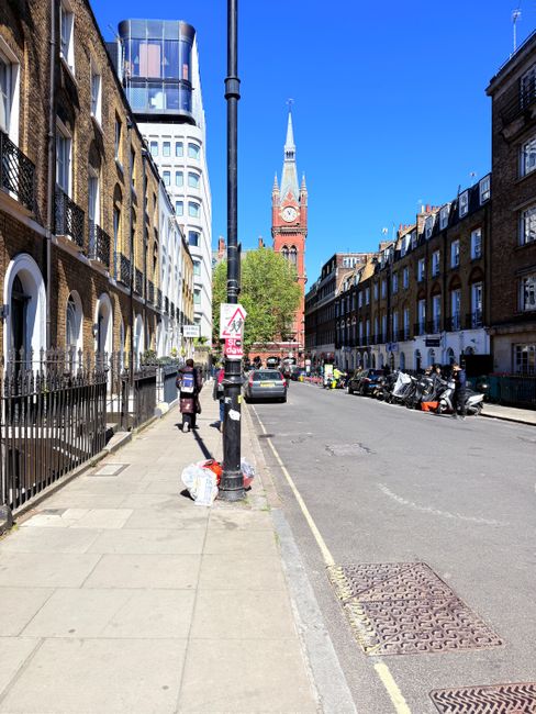 A city street scene featuring a paved sidewalk with a metal railing on the left side and a row of terraced brick residential buildings with white window frames. The sidewalk has some garbage bags plac