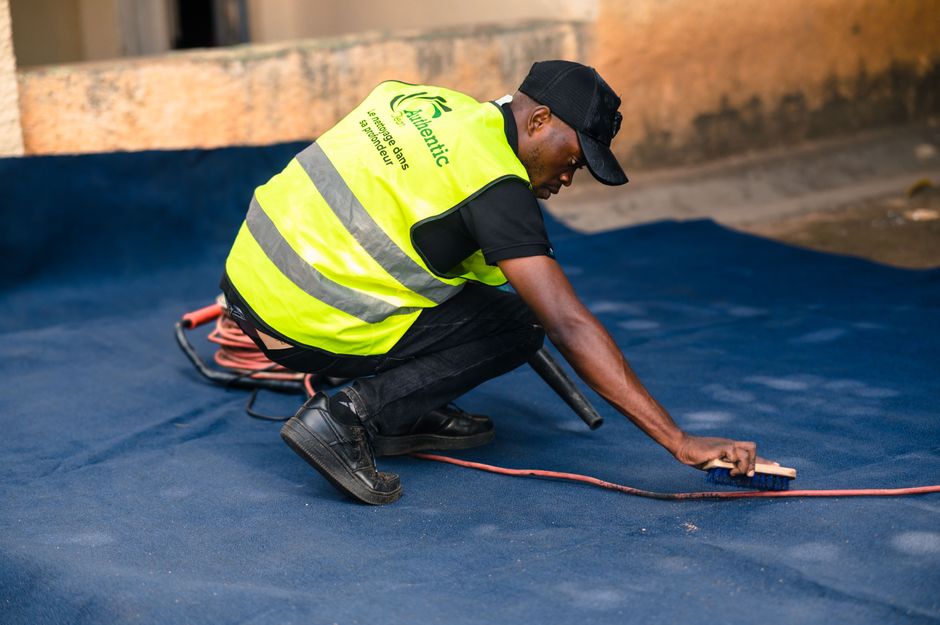 A person dressed in orange work overalls using a yellow wet vacuum cleaner with a black hose attachment to perform surface cleaning on a tiled floor inside a modern living room. The vacuum cleaner is 